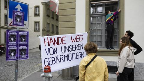Ein Banner mit der Aufschrift "Hände weg von unseren Gebärmüttern" wird aufgehängt, mehrere Menschen schauen zu.