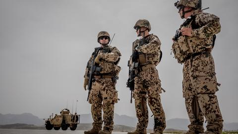Drei Soldaten im Tarnanzug mit Gewehr vor einem Panzer in Wüstenlandschaft