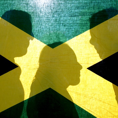 Eine Frau und zwei Männer stehen als Schattenriss sichtbar hinter der jamaikanischen Landesfahne mit den Farben Schwarz, Gelb und Grün
