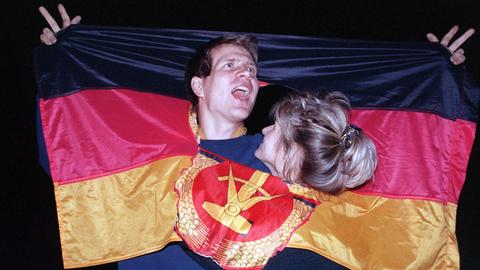 ARCHIV - 03.10.1990, Berlin: Ein jubelnden Paar hat sich am Tag der Deutschen Einheit in einer DDR-Fahne dort Platz geschaffen, wo früher das Emblem Hammer und Sichel war - und feierte frenetisch den historischen Anlaß.