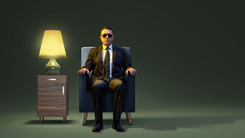 Ein herrischer Mann mit Sonnenbrille sitzt auf einem Sessel einem Kulissenraum