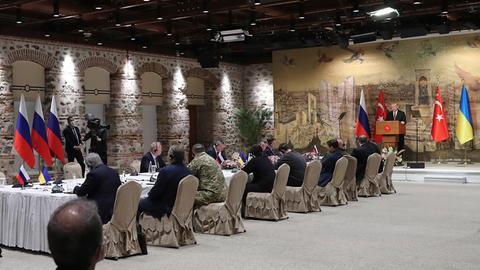 Männer sitzen an einem langen Tisch, im Hintergrund ein Redner vor Flaggen der Türkei, Russlands und der Ukraine