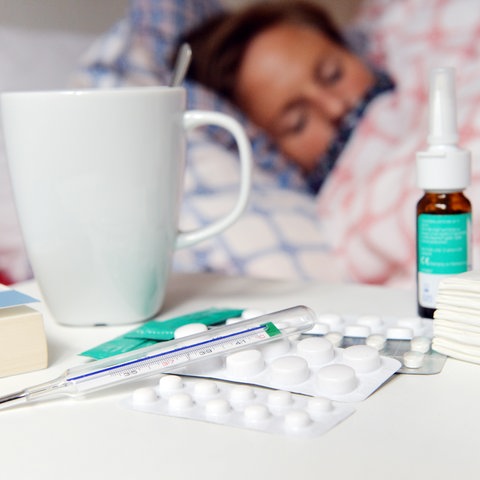 Medikamente und ein Fiberthermometer liegen im Vordergrund während man im Hintergrund einen an Grippe erkrankten Patienten im Bett liegen sieht.