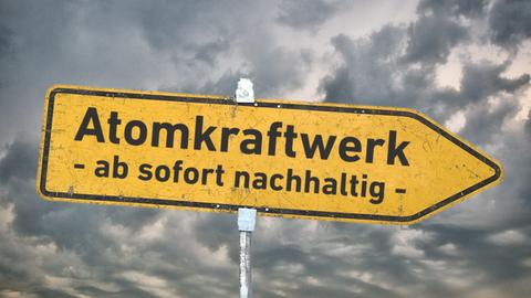 Ein gelbes, zerkratztes Hinweisschild mit der Aufschrift "Atomkraftwerk - ab sofort nachhaltig -" vor einem grau bewölkten Himmel mit aufziehendem Gewitter. 