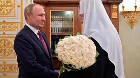 Ein Mann im Anzug schüttelt einem orthodoxen Bischof mit weißer Haube und einem Strauß Rosen die Hand.