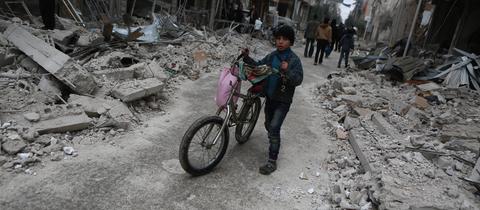 Syrien, Ghuta: Ein Kind schiebt sein Fahrrad an Gebäuden vorbei, die durch Angriffe der syrischen Luftwaffe zerstört wurden.