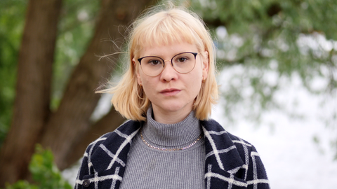 Blonde junge Frau mit Brille vor Bäumen.