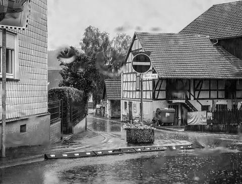 Impressionen aus dem Vogelsberg: Im Regen durch eine Scheibe fotografierte Dorfstrassenkreuzung mit Verkehrsinsel vor Fachwerkhaus