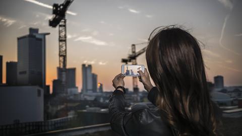 junge Frau fotografiert mit ihrem Mobilfunkgerät die Frankfurter Skyline vor ABendhimmel 