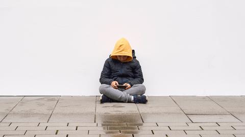 Ein Junge mit gelbem Kapuzenpulli und blauer Jacke sitzt auf dem Boden, gebeugt über sein Mobilfunkgerät, mit dem er beschäftigt ist. 