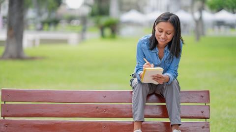 Eine junge Latina sitzt auf Rückenlehne einer Parkbank und schreibt in eine Heft