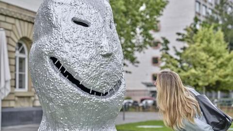 Eine Frau schaut eine große grimmig schauende Figur aus Ugo Rondinones Kunstwerk "Sunrise.East“ an (Ausstellung im Garten des Städel Museums)