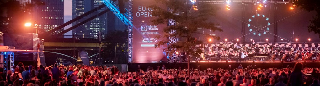 Im Dunkeln rot-lila beleuchtete Bühne mit Orchester und Publikum davor. Im Hintergrund die Frankfurter Skyline