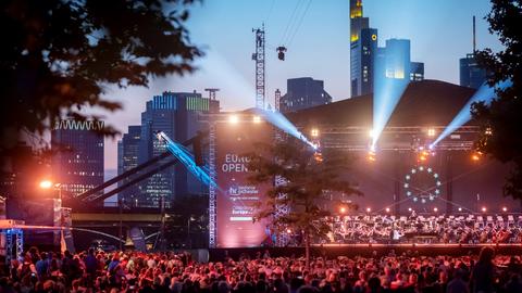 Im Dunkeln rot-lila beleuchtete Bühne mit Orchester und Publikum davor. Im Hintergrund die Frankfurter Skyline